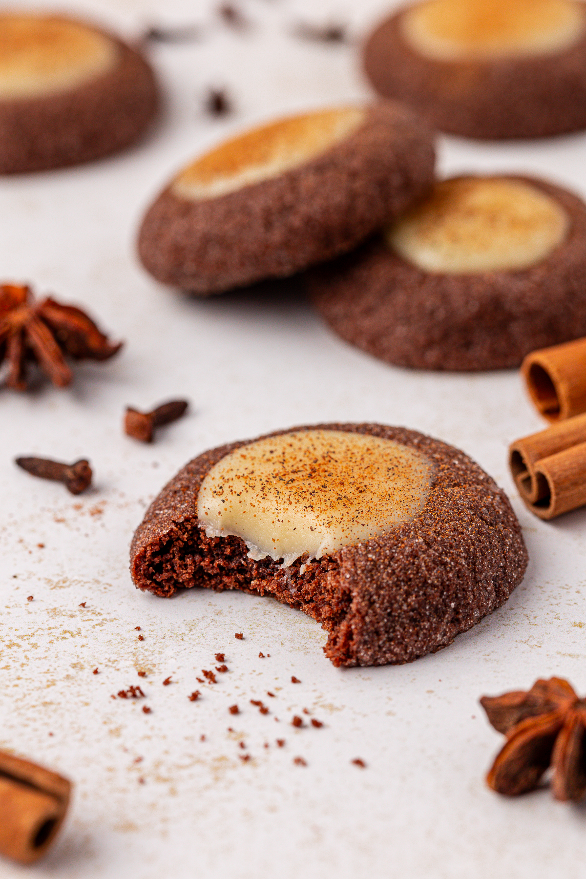 A close-up of a chocolate cookie with a creamy center and a bite taken out, surrounded by whole cookies, cinnamon sticks, and star anise on a white surface.