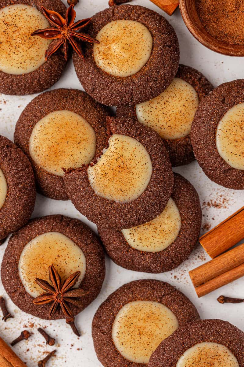 A close-up of chocolate cookies with creamy centers, sprinkled with cinnamon, surrounded by cinnamon sticks, star anise, and cloves on a white surface. One cookie has a bite taken out of it.
