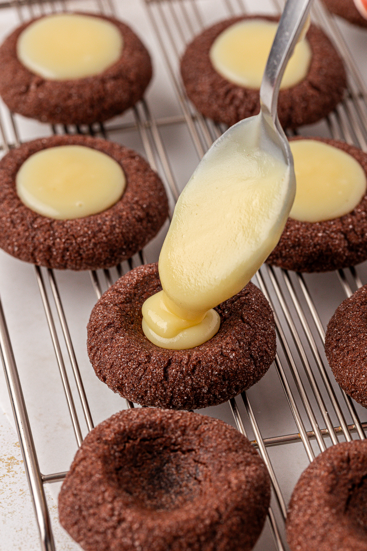 A spoon is filling the center of a chocolate thumbprint cookie with creamy yellow filling on a wire cooling rack, surrounded by other similar cookies.