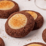 Close-up of chocolate thumbprint cookies with a creamy, light-colored filling in the center, sprinkled with cinnamon or cocoa powder, arranged on a white surface.
