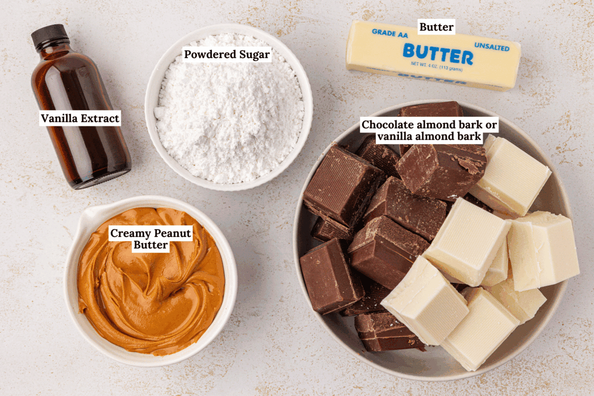Labeled ingredients for a recipe arranged on a countertop: a bottle of vanilla extract, a bowl of powdered sugar, a bowl of creamy peanut butter, a stick of unsalted butter, and a plate with chocolate and white almond bark pieces.