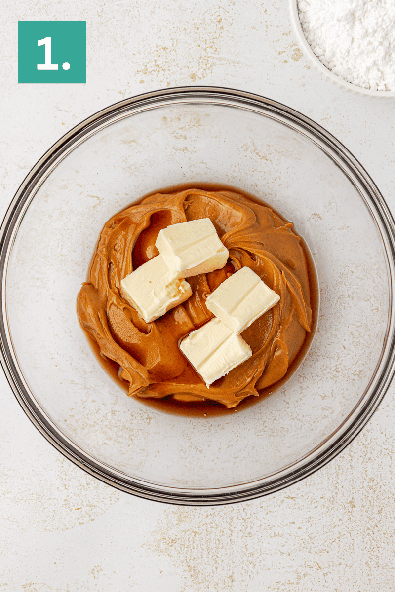 A glass mixing bowl with creamy peanut butter, vanilla extract, and chunks of butter inside. A green box with the number 1 is in the top left corner, and a bowl of powdered sugar is in the top right.