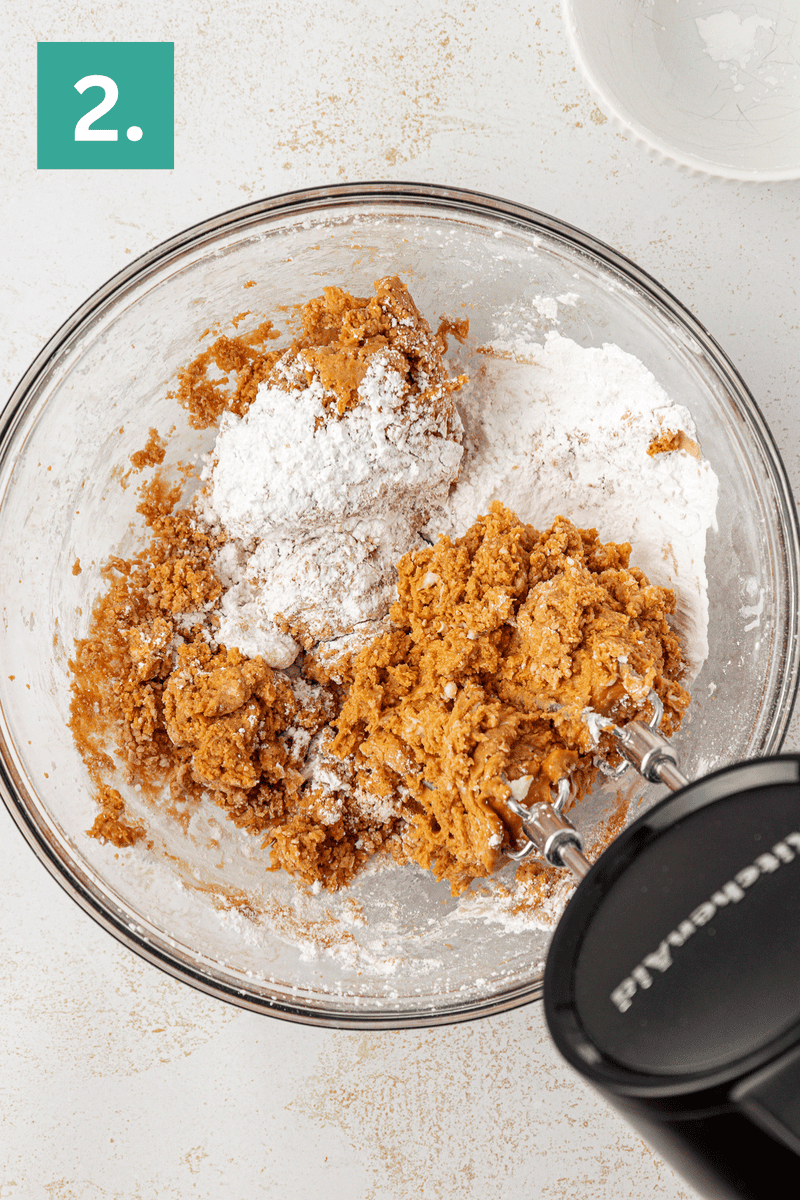 A glass bowl with brown cookie dough and white flour being mixed with an electric hand mixer, numbered step 2 in the top left corner. A white bowl is partially visible in the upper right background.