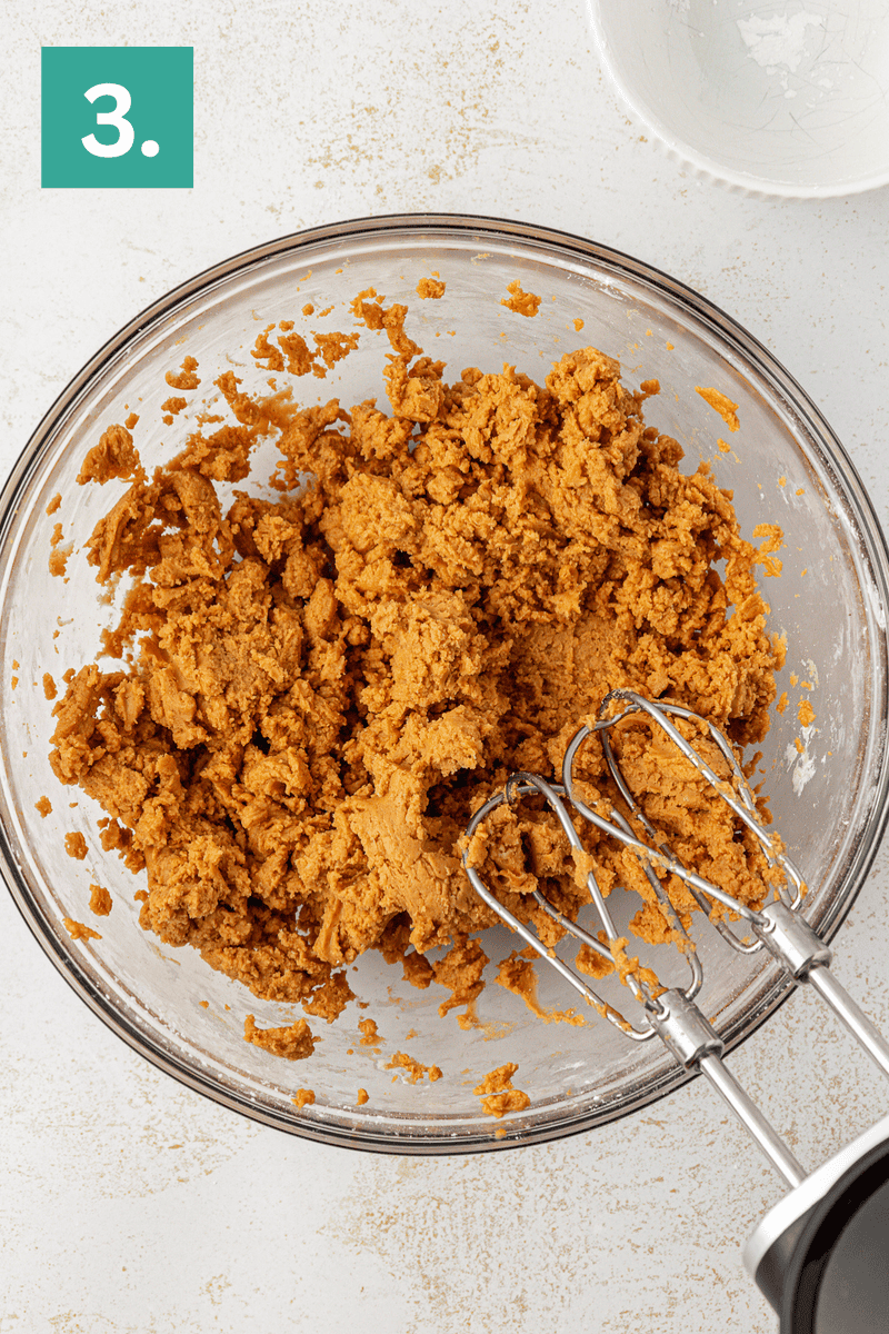 A glass bowl filled with brown cookie dough and a hand mixer with beaters, shown from above. A green box with the number 3 is in the top left corner, and a white bowl sits nearby on a white surface.