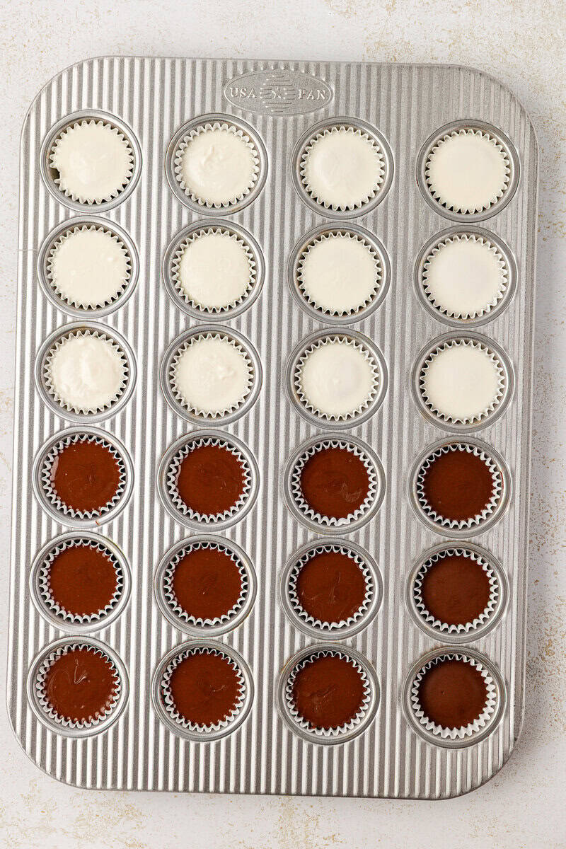 A muffin tin with 24 cups; the top two rows are filled with white batter, and the bottom two rows are filled with chocolate batter, each in a white cupcake liner.