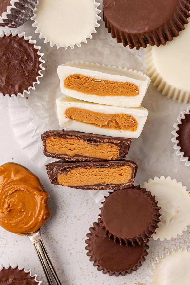 An assortment of chocolate and white chocolate peanut butter cups, some whole and some cut in half, are arranged on a light surface with empty paper wrappers and a spoonful of peanut butter nearby.