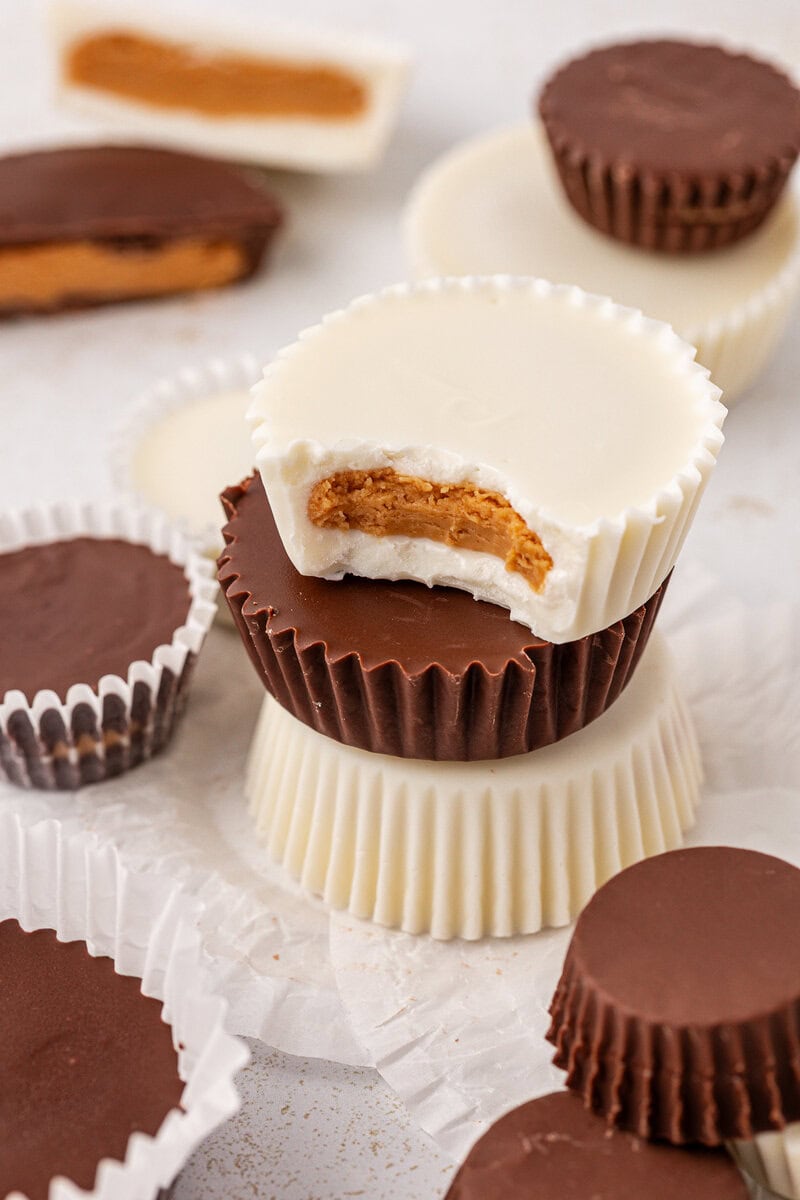 A stack of chocolate and white chocolate peanut butter cups, with one white chocolate cup on top showing a bite revealing the peanut butter filling inside. Several more cups are scattered around on paper cupcake liners.