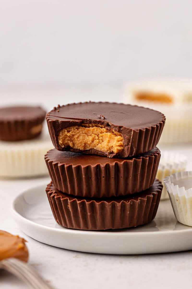 Three chocolate peanut butter cups are stacked on a plate; the top one has a bite taken out, revealing a creamy peanut butter filling. In the background, more peanut butter cups and paper wrappers are visible.