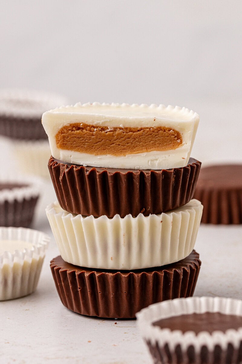 A stack of chocolate and white chocolate peanut butter cups, with the top white chocolate cup cut in half to show the peanut butter filling inside. Other peanut butter cups are blurred in the background.