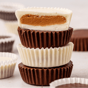 A stack of chocolate and white chocolate peanut butter cups sits on a light surface, with the top cup sliced in half to reveal the creamy peanut butter filling inside. Other cups are blurred in the background.
