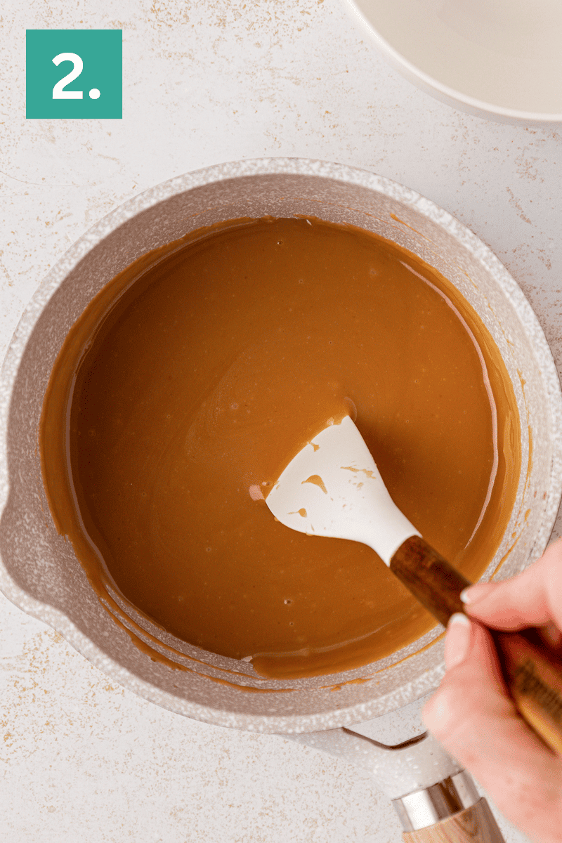 A hand stirs smooth caramel sauce with a white spatula in a light-colored saucepan. A green box with the number 2 is in the upper left corner.