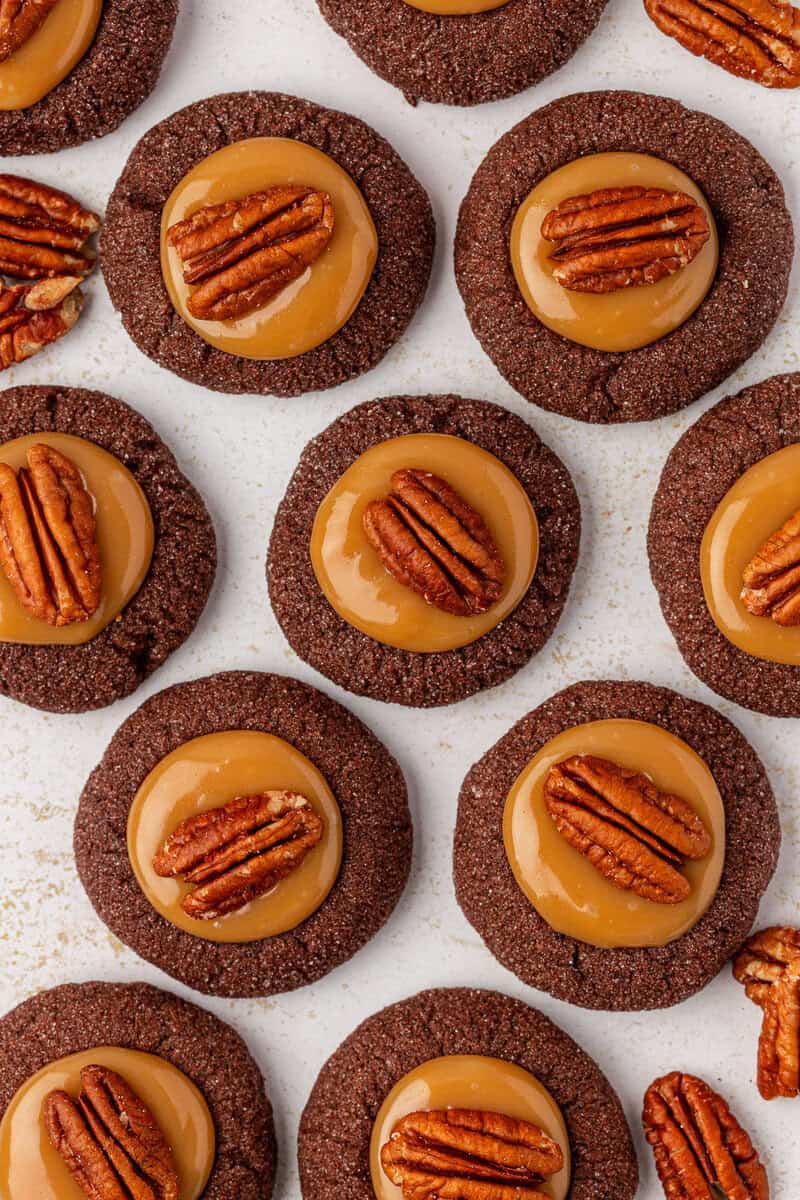 Chocolate cookies topped with caramel and a pecan half are arranged in neat rows on a light surface, creating a uniform and appetizing pattern.