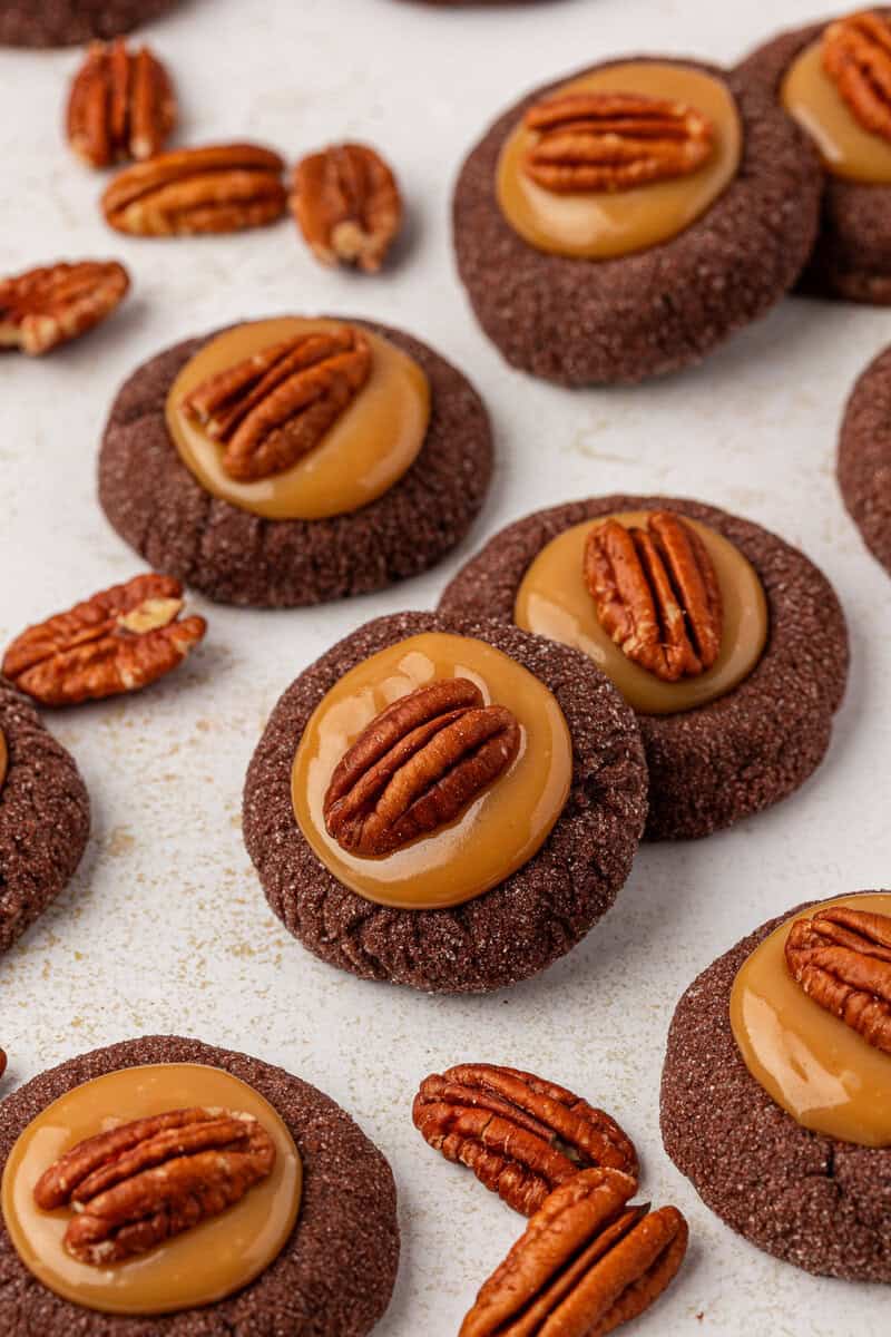Round chocolate cookies topped with a glossy caramel layer and a whole pecan, scattered on a white surface with some loose pecans around them.