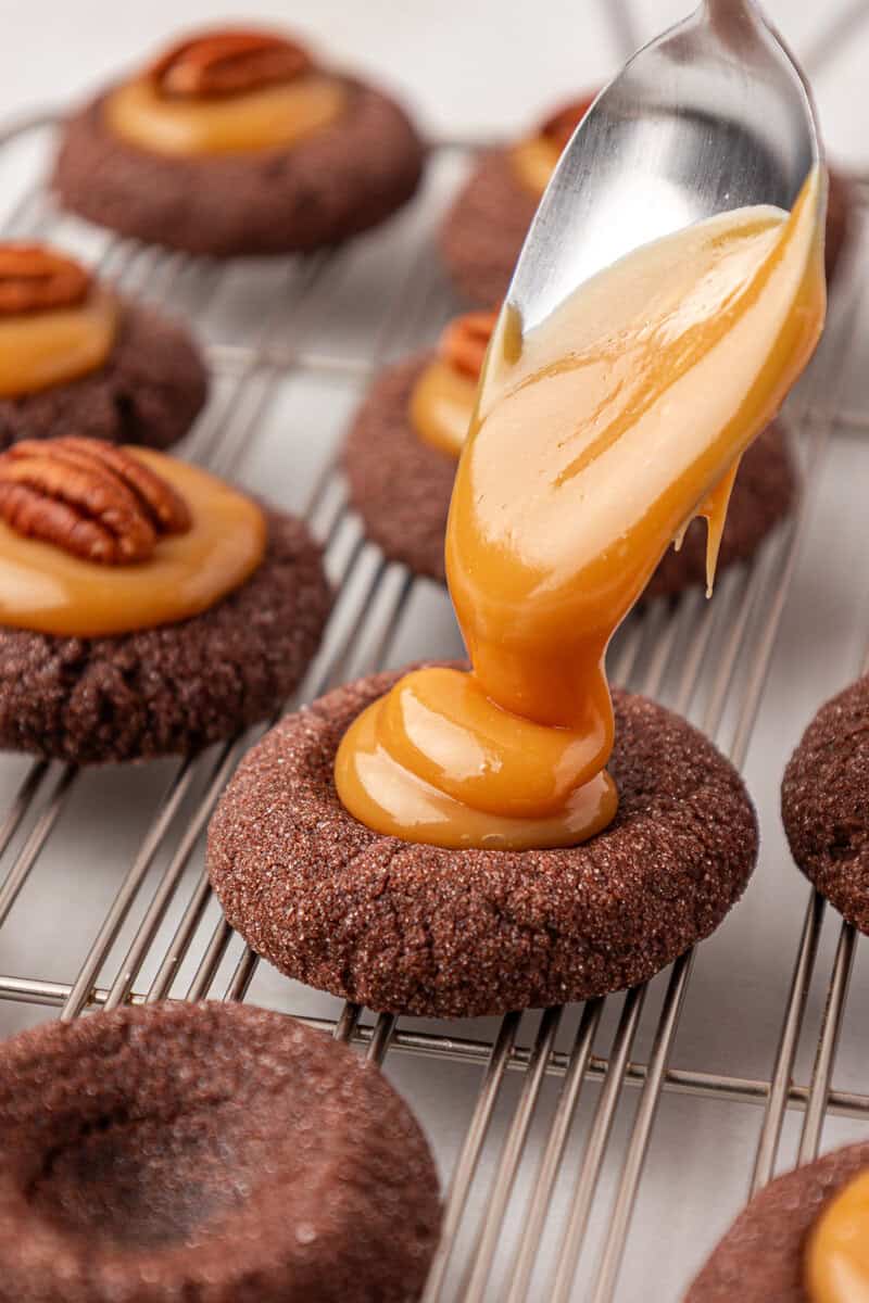 A spoon drizzles caramel onto a round chocolate cookie topped with a pecan, while similar cookies cool on a wire rack in the background.
