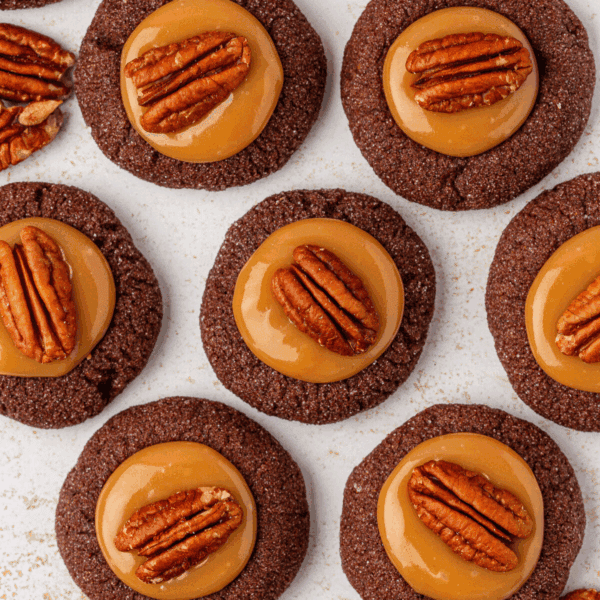 Chocolate cookies topped with a layer of caramel and a pecan half, arranged neatly on a white surface. The cookies have a glossy caramel center, and the pecans add a decorative touch.