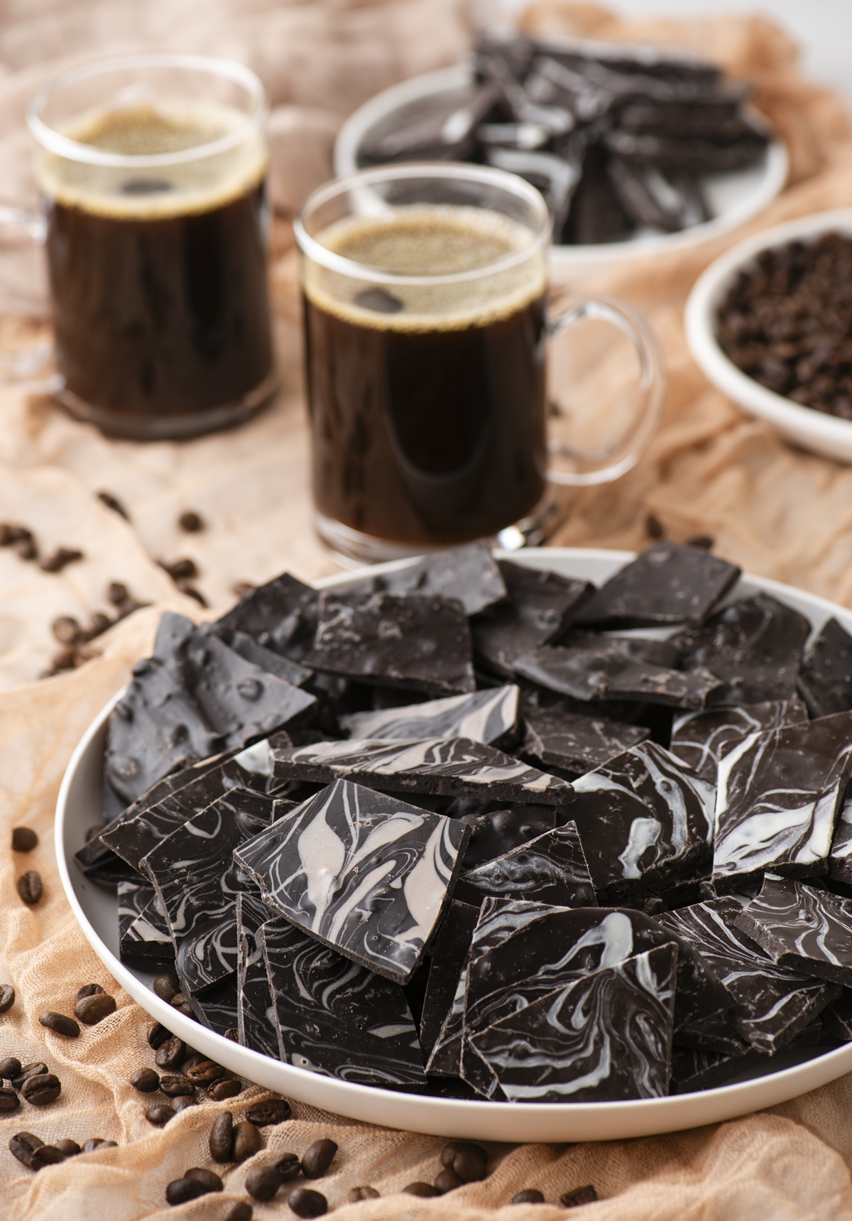 A plate of marbled dark chocolate pieces sits on a fabric-covered surface, surrounded by scattered coffee beans. Two glass mugs of black coffee and more chocolate pieces are in the background.