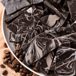 A plate of homemade coffee bean chocolate bark, some with a marbled white and dark chocolate pattern, surrounded by whole coffee beans. Text on the image reads: No Bake Recipe - Coffee Bean Bark - thefirstyearblog.com.