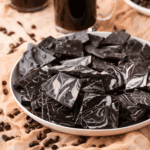 A white plate filled with pieces of dark chocolate with white swirls sits on a beige cloth, surrounded by scattered coffee beans and a glass mug in the background.