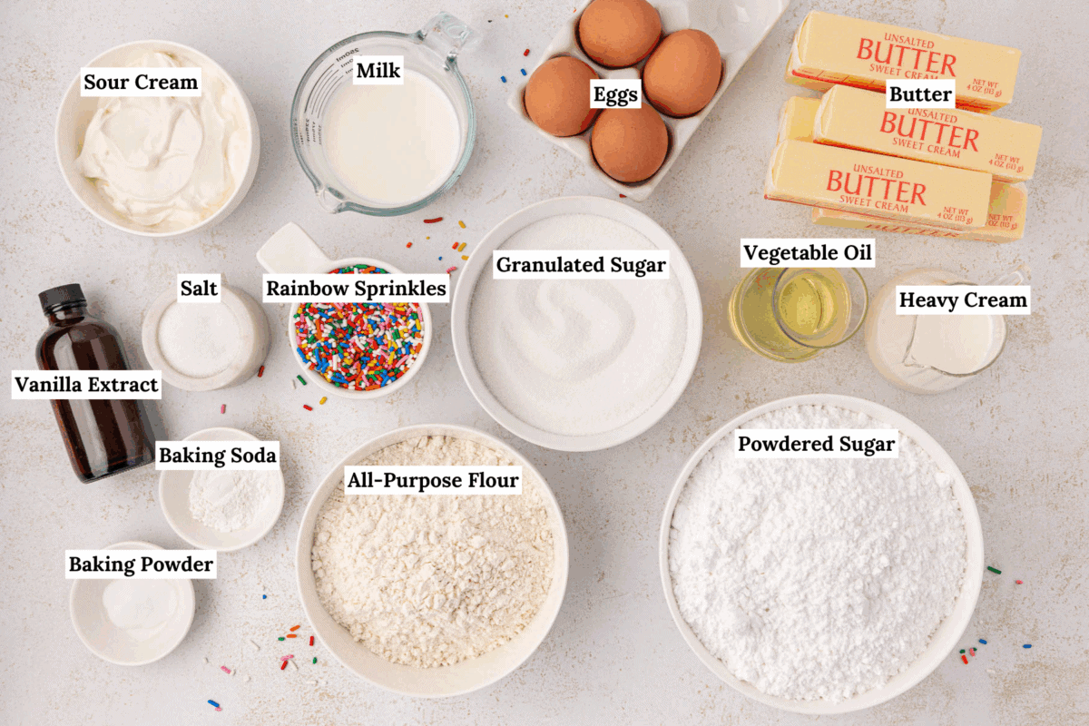 Top-down view of labeled baking ingredients on a white surface, including eggs, butter, milk, heavy cream, vegetable oil, sour cream, rainbow sprinkles, vanilla extract, salt, sugars, flour, baking powder, and baking soda.