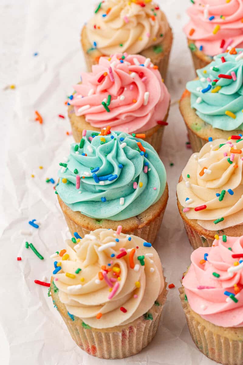 A group of cupcakes with colorful swirled frosting in pastel pink, blue, and cream, topped with rainbow sprinkles, arranged on crumpled white parchment paper.