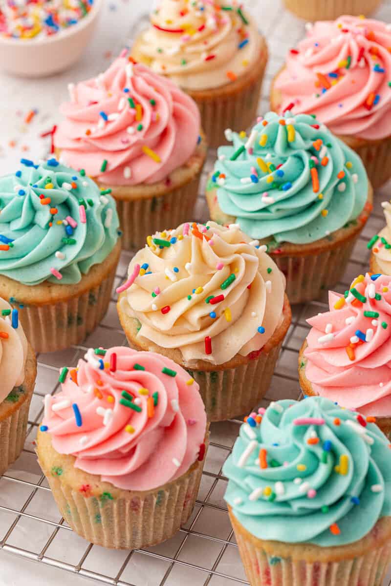 Colorful cupcakes with swirled pink, blue, and cream frosting are topped with rainbow sprinkles and arranged on a cooling rack. A small bowl of extra sprinkles is visible in the background.