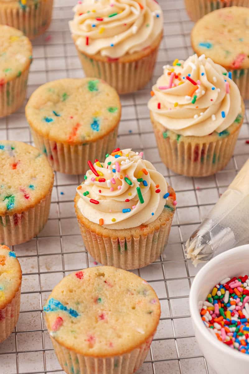 Funfetti cupcakes on a wire cooling rack, some topped with swirls of vanilla frosting and colorful sprinkles, others plain. A bowl of rainbow sprinkles and a piping bag are nearby.