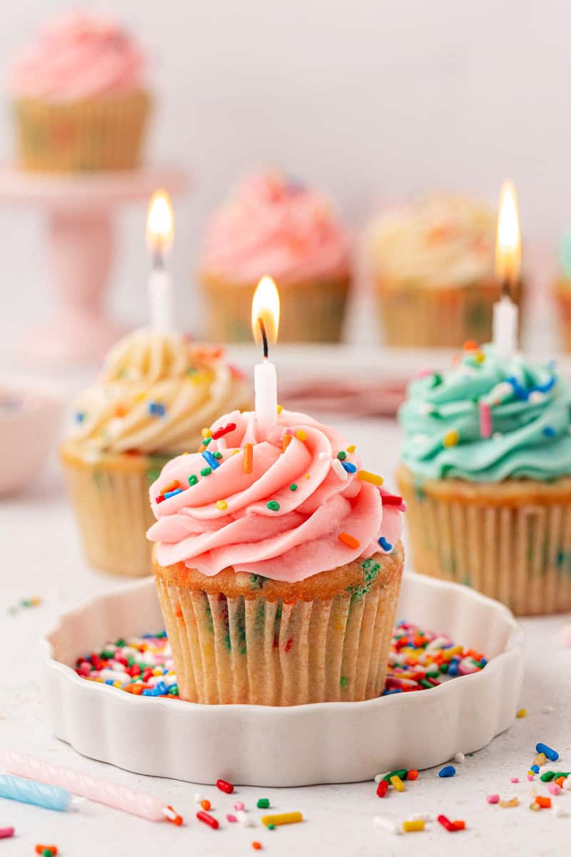 A cupcake with pink frosting, colorful sprinkles, and a lit candle sits on a plate surrounded by more sprinkles. Other cupcakes with different frosting colors and lit candles are blurred in the background.