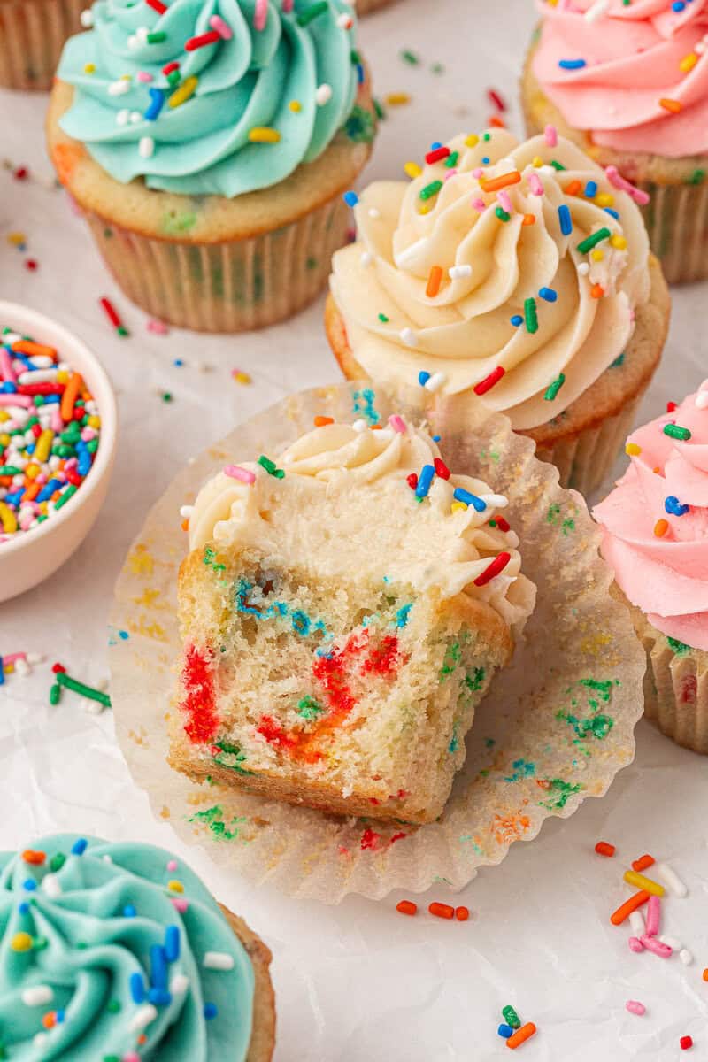 Colorful cupcakes with swirled frosting and rainbow sprinkles are displayed, with one cupcake unwrapped and partially eaten, revealing a funfetti interior. A small bowl of sprinkles sits nearby on a white background.
