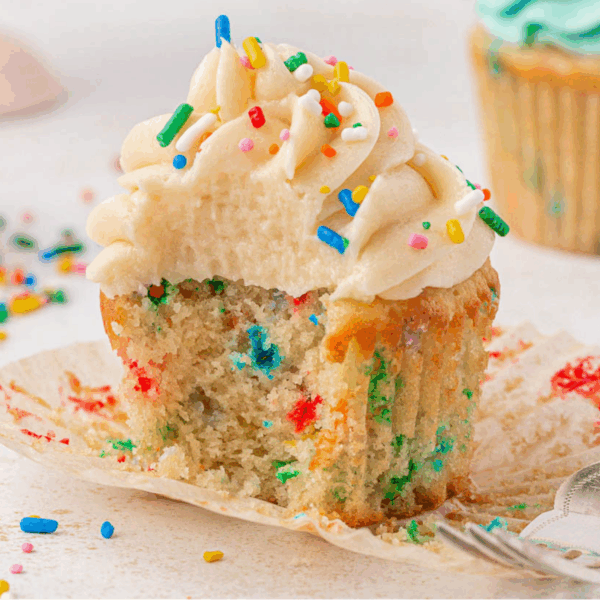 A partially unwrapped cupcake with vanilla frosting and colorful sprinkles, showing a moist, confetti-filled interior. Some sprinkles are scattered on the surface around the cupcake.