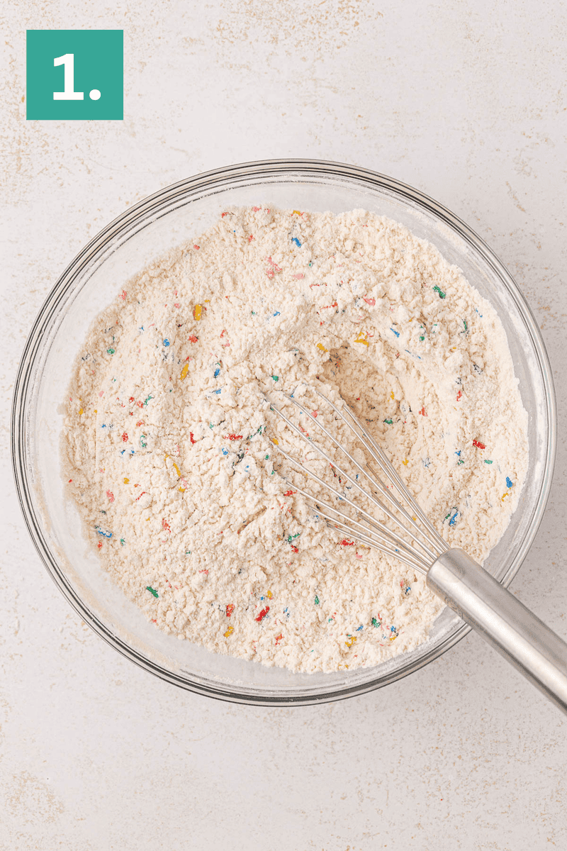 A glass bowl filled with flour mixed with colorful sprinkles and a metal whisk, shown on a light countertop. A green box with the number 1 is in the top left corner.