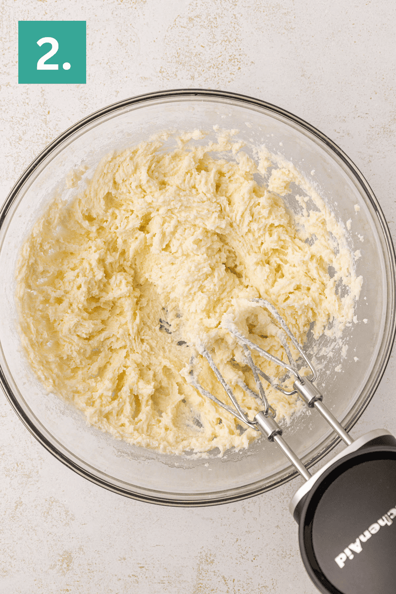 A glass mixing bowl with creamed butter and sugar sits on a light surface. An electric hand mixer with beaters is resting in the bowl. A green square in the corner displays the number 2.