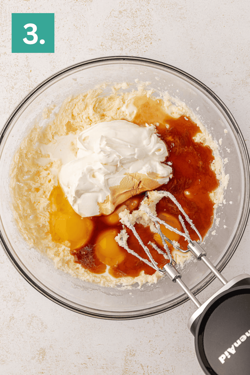A glass mixing bowl with eggs, sour cream, vanilla, and other ingredients on top of a creamy mixture. An electric hand mixer with beaters is resting in the bowl. A green box with the number 3 is in the corner.