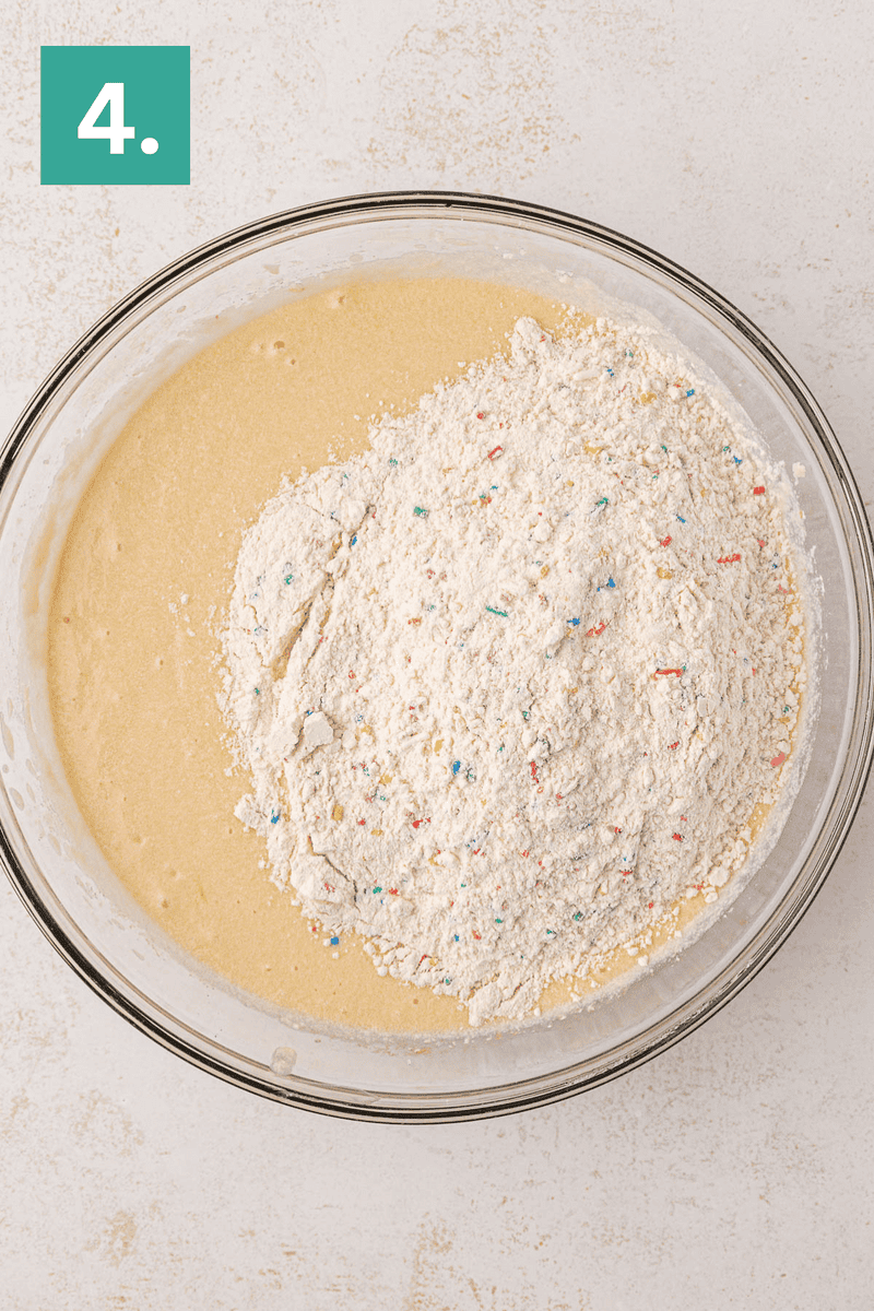 A large glass bowl containing cake batter with a mound of flour and colorful sprinkles on top. A green square with the number 4 is in the top left corner.