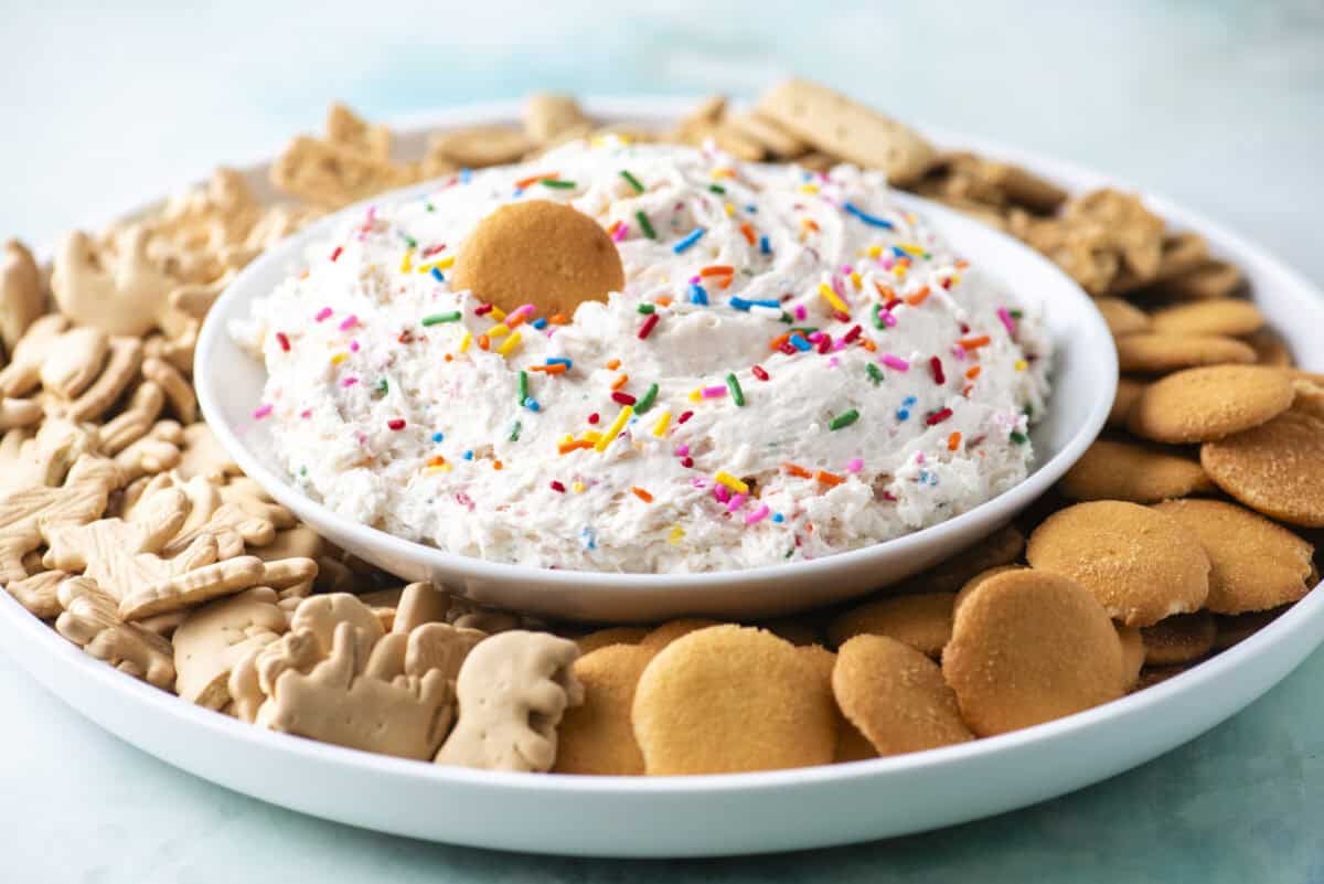 A plate with a bowl of funfetti dip topped with rainbow sprinkles and a vanilla wafer, surrounded by animal crackers, graham crackers, and vanilla wafer cookies.