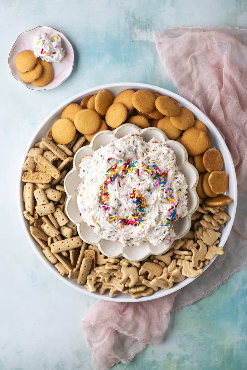 A large tray with a bowl of funfetti dip topped with rainbow sprinkles, surrounded by vanilla wafers, graham crackers, and animal crackers, with a small plate of cookies and dip on the side.