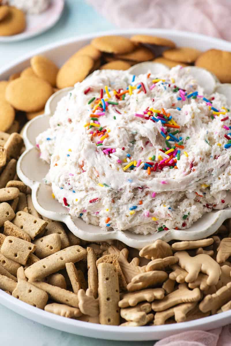 A plate of fluffy dip topped with colorful sprinkles is surrounded by vanilla wafers, animal crackers, and rectangular cookies on a large serving platter.