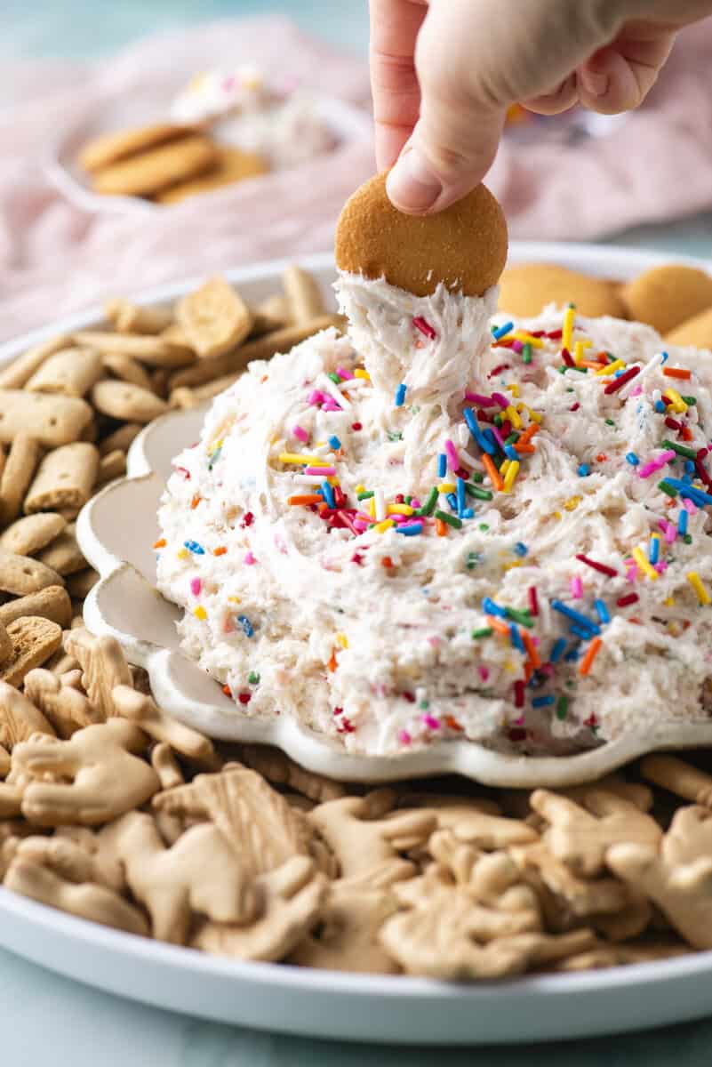 A hand dips a round cookie into a fluffy, sprinkle-topped dip on a plate surrounded by animal crackers and cookies. The colorful sprinkles decorate the creamy dessert.