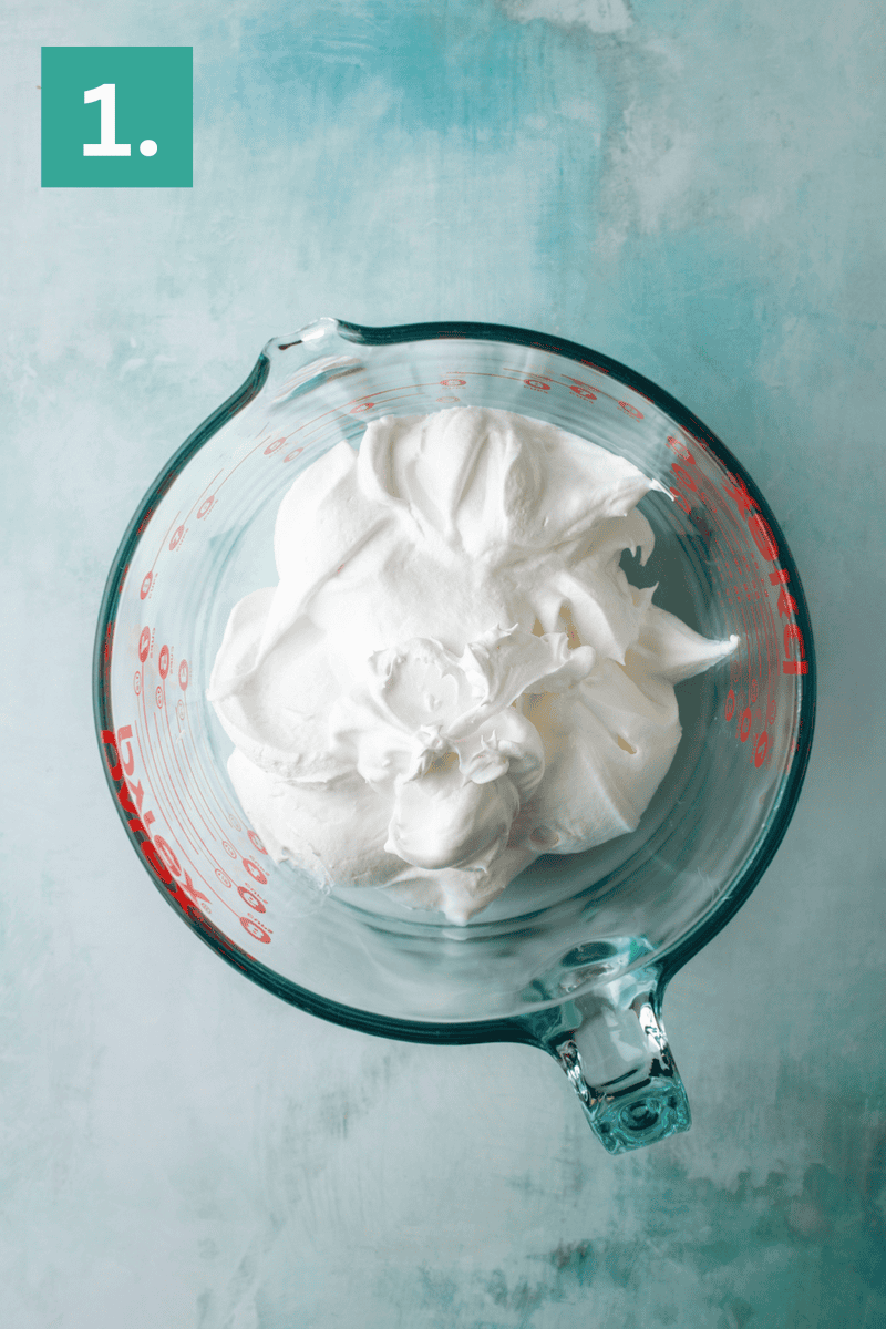 A glass measuring cup filled with whipped cream sits on a light blue surface. A green box with the number 1 is in the top left corner of the image.