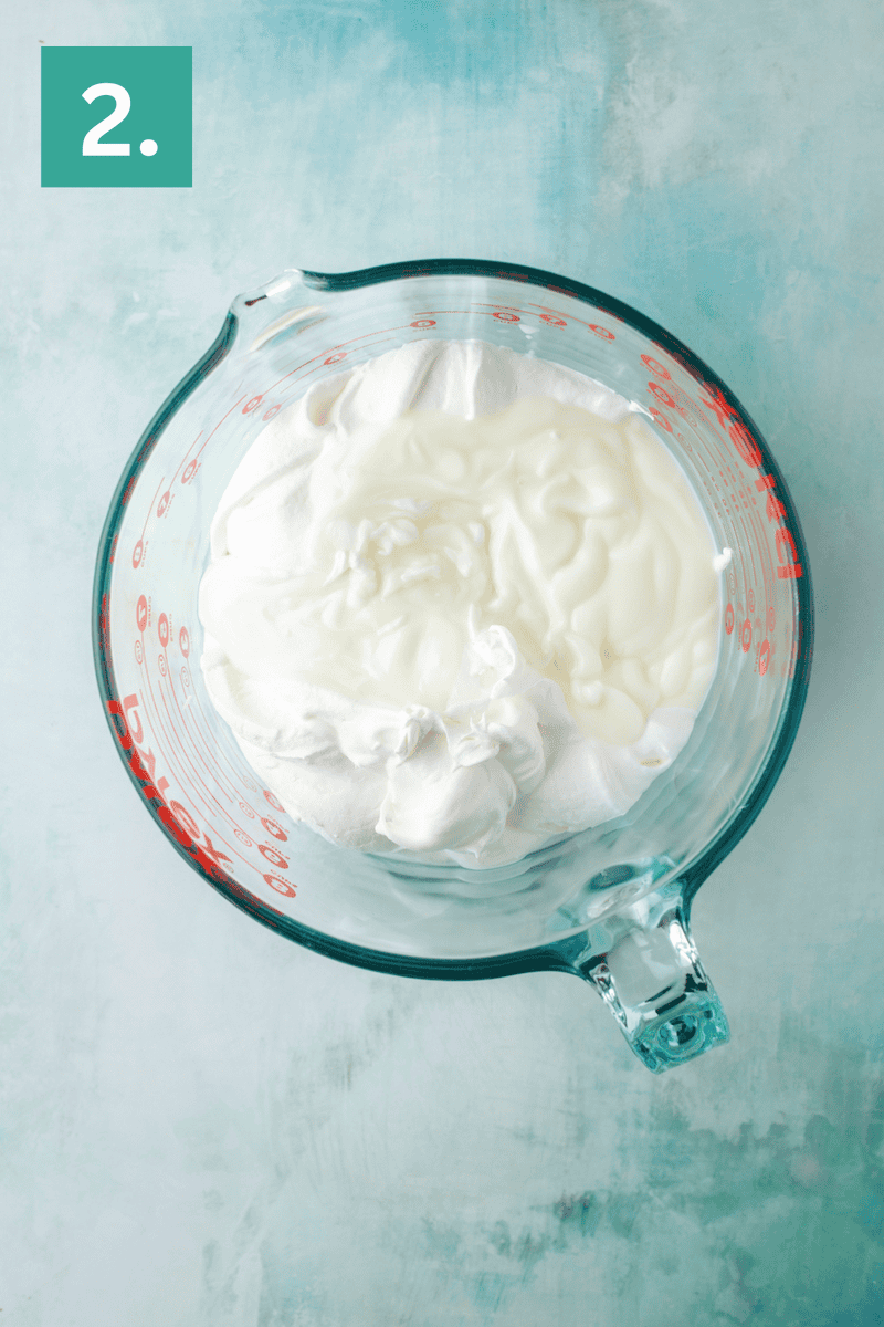 A glass measuring cup filled with creamy white ingredients, likely yogurt and whipped topping, sits on a light blue surface. A green box with the number 2. is in the top left corner.