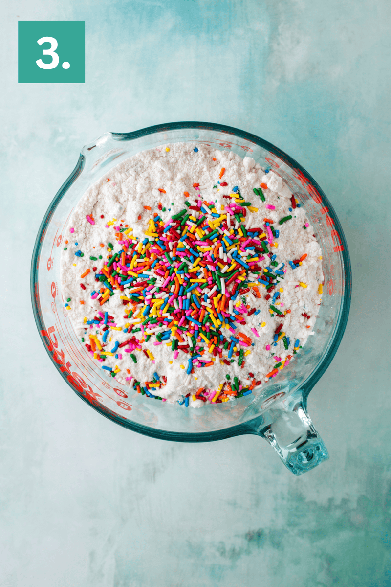 A glass measuring cup filled with flour and colorful rainbow sprinkles sits on a light blue surface. A green box with the number 3 is in the upper left corner.