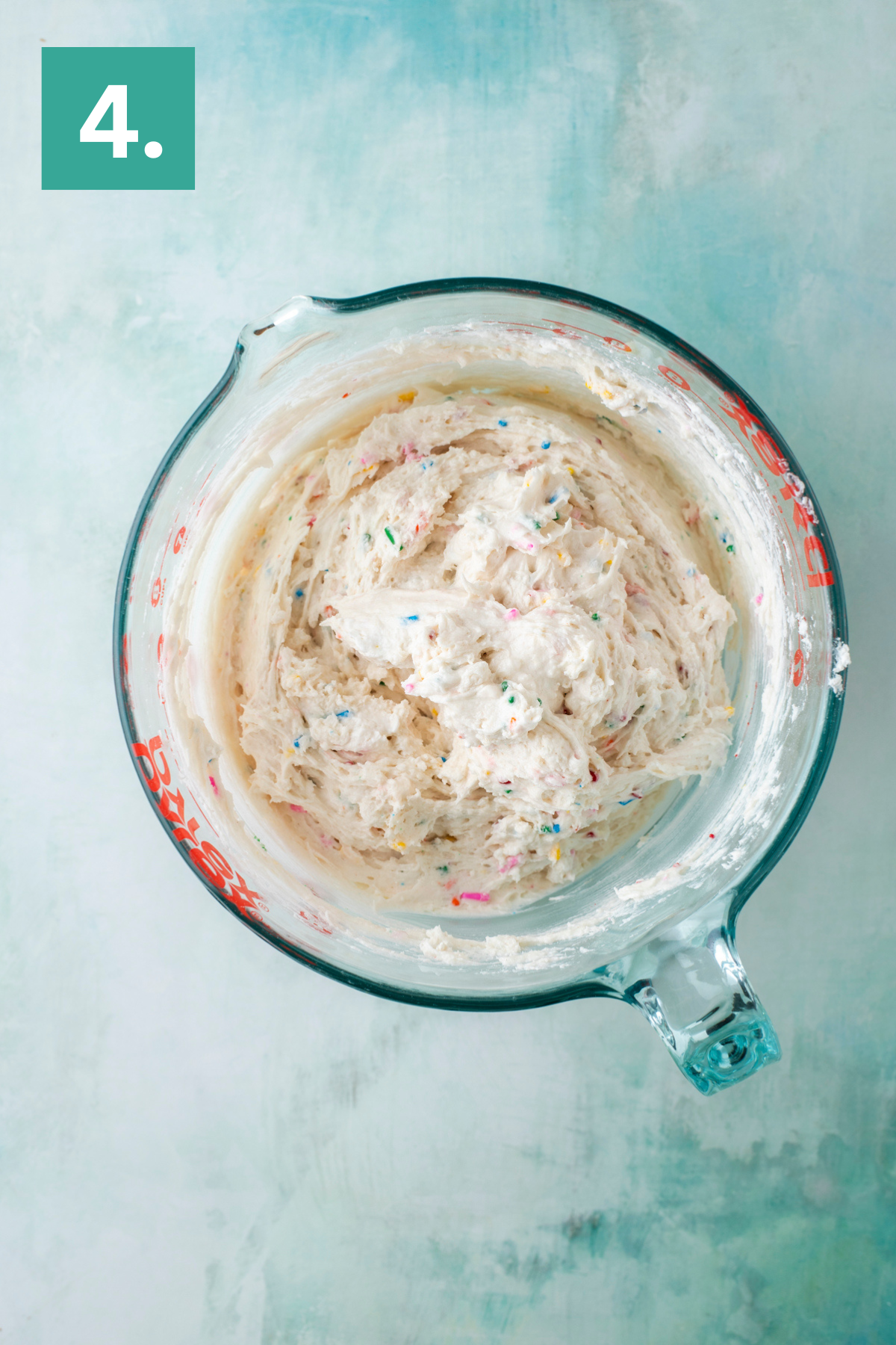 A glass mixing bowl filled with creamy, funfetti cake batter sits on a light blue surface. A green box with the number 4 is in the top left corner, indicating step four of a recipe.