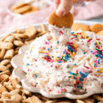 A hand dips a round cookie into a creamy, sprinkle-topped dip surrounded by various cookies and animal crackers on a decorative plate.