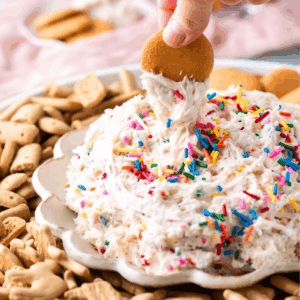 A hand dips a round cookie into a creamy, sprinkle-topped dip surrounded by various cookies and animal crackers on a decorative plate.