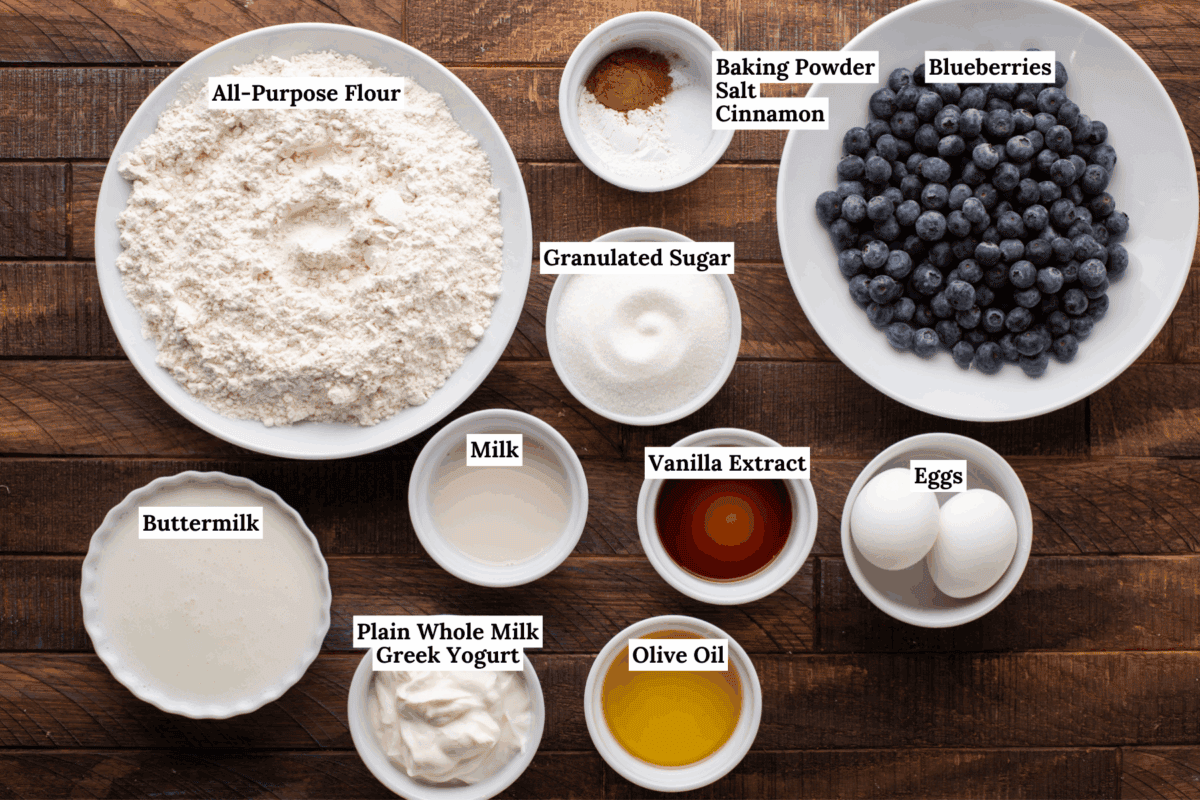 Various baking ingredients in bowls on a wooden surface, including all-purpose flour, granulated sugar, buttermilk, milk, Greek yogurt, olive oil, vanilla extract, eggs, blueberries, and a mix of baking powder, salt, and cinnamon.