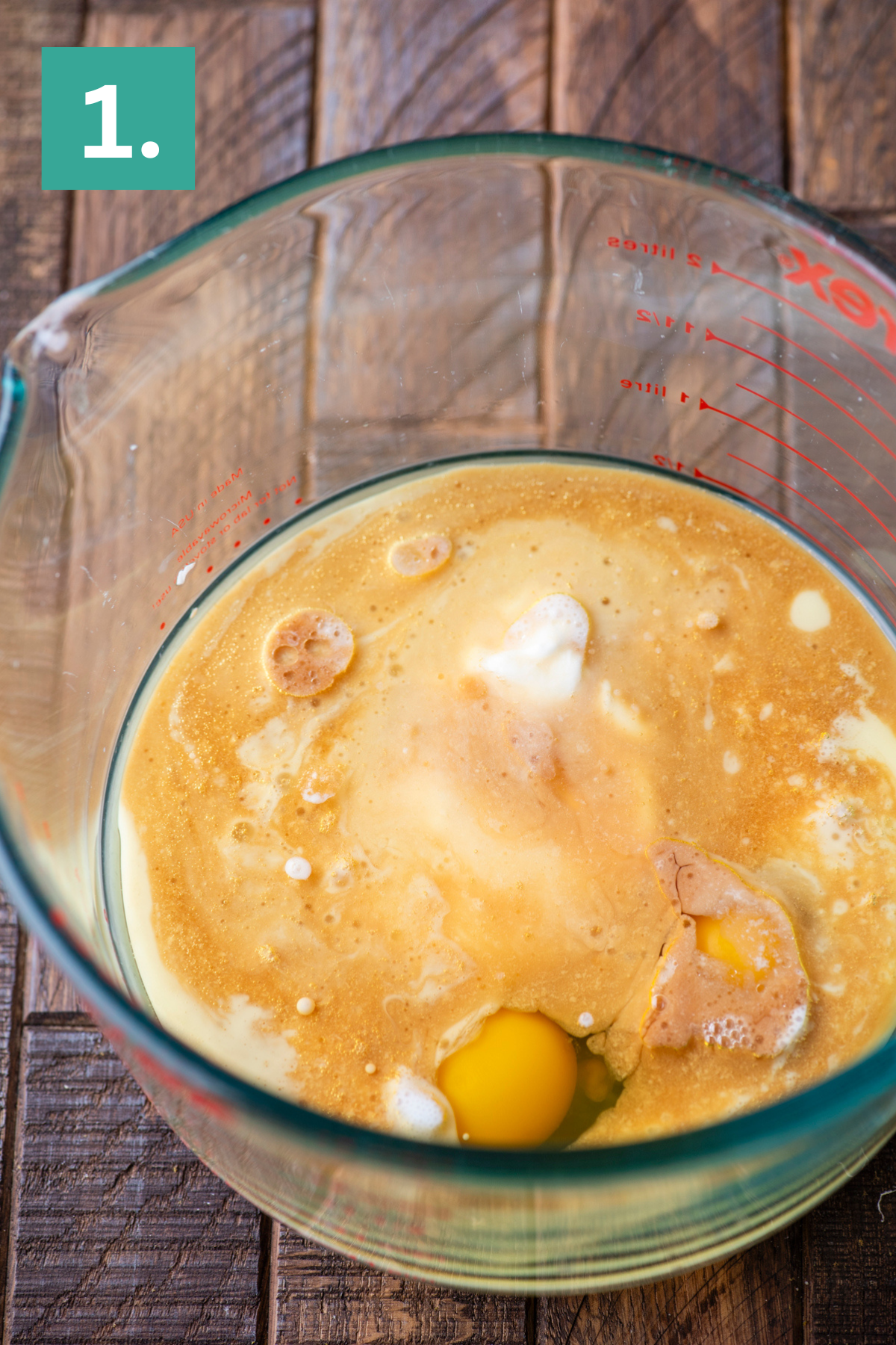 A large glass measuring cup on a wooden surface contains milk, eggs, and other liquid baking ingredients. A green box with the number 1. appears in the top left corner, indicating the first step of a recipe.