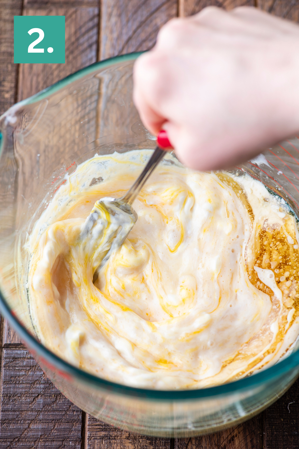 A hand stirs creamy ingredients with a metal fork in a glass bowl on a wooden surface. A green square with the number 2 is in the upper left corner.