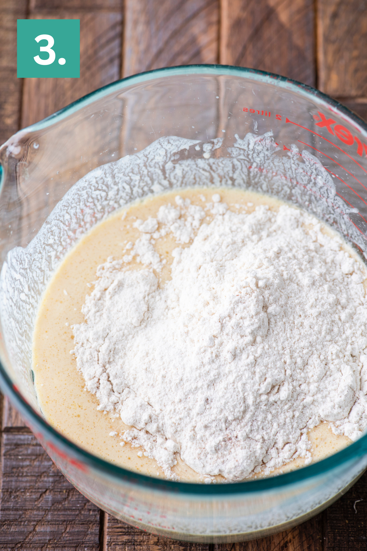 A glass measuring bowl with a mixture of flour and wet ingredients sits on a wooden surface. A green box labeled 3. is in the top left corner, indicating this is step three in a recipe.