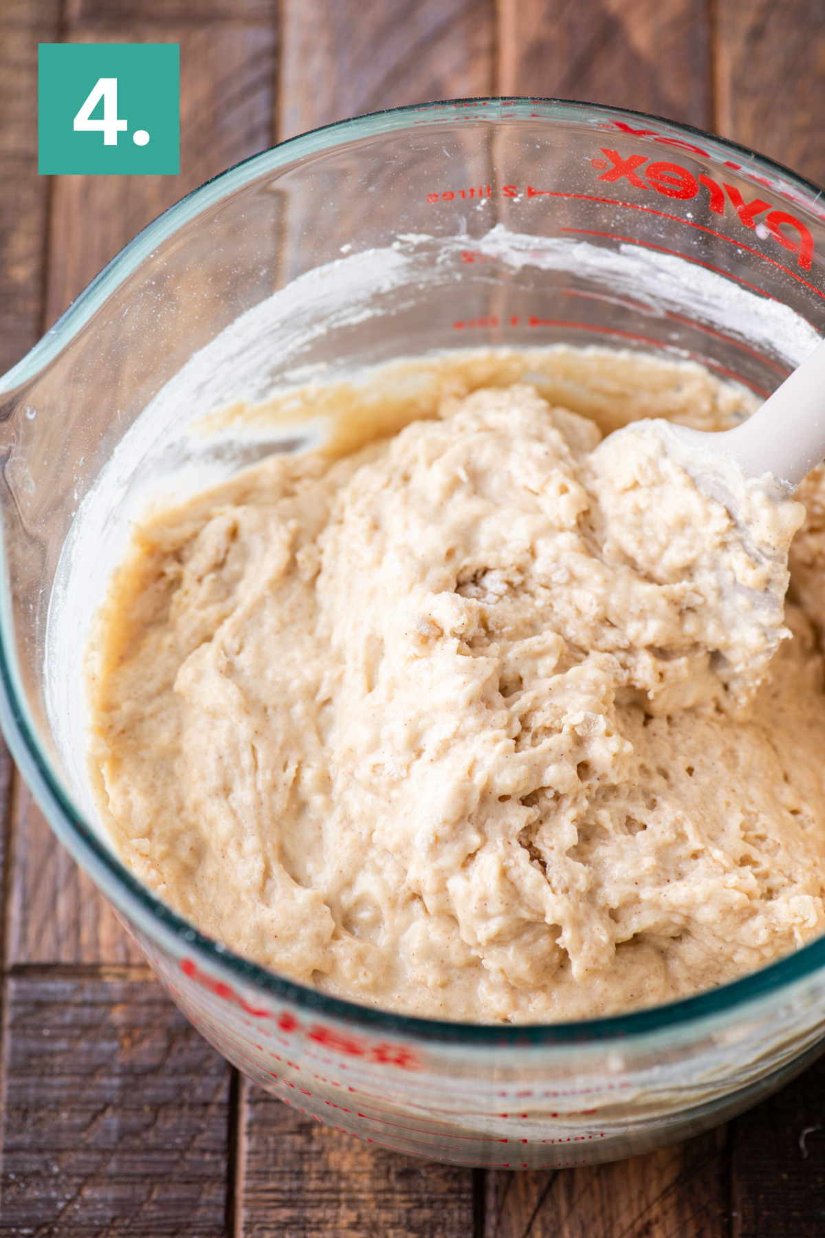 A glass measuring bowl with thick bread dough being mixed with a white spatula, resting on a wooden surface. A green square with the number 4 is in the top left corner.