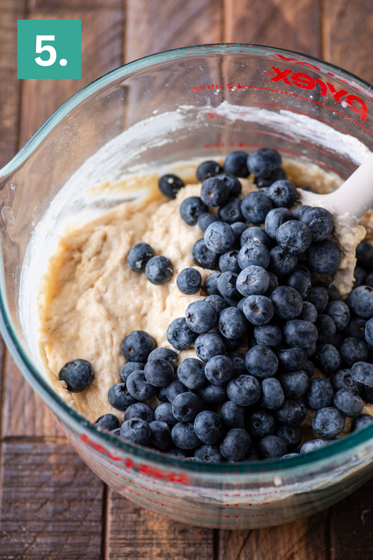 A glass mixing bowl filled with muffin batter and fresh blueberries, being stirred with a white spatula, sits on a wooden surface. A green box with the number 5 appears in the top left corner.