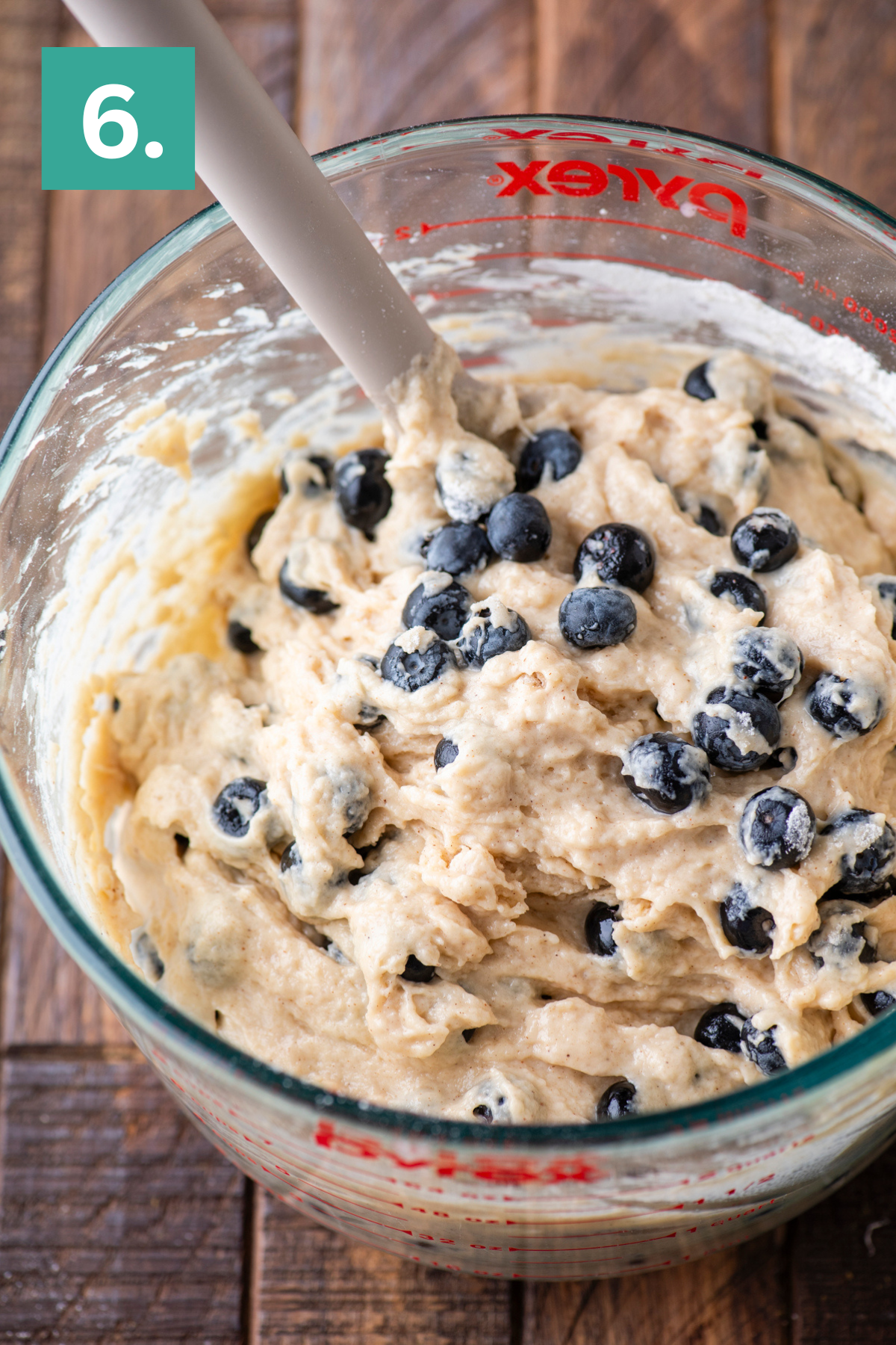 A glass mixing bowl filled with thick blueberry muffin batter and a spatula. The bowl sits on a wooden surface, and a green square with the number 6 is in the top left corner.